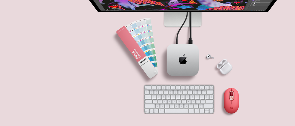 Bovenkant van Mac mini, zilverkleurig aluminium, afgeronde hoeken, glanzend zwart Apple logo in het midden, verbonden met een kabel, Studio Display deels zichtbaar aan de bovenkant, AirPods 4, opengeklapt, Pantone-kleurstalen, en Logitech-toetsenbord en -muis in rode kleur